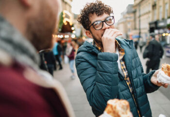 man wearing winter clothes and eating a sandwich while talking to a friend