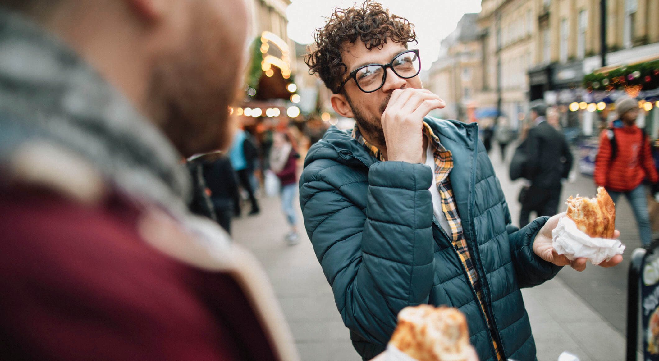 man wearing winter clothes and eating a sandwich while talking to a friend