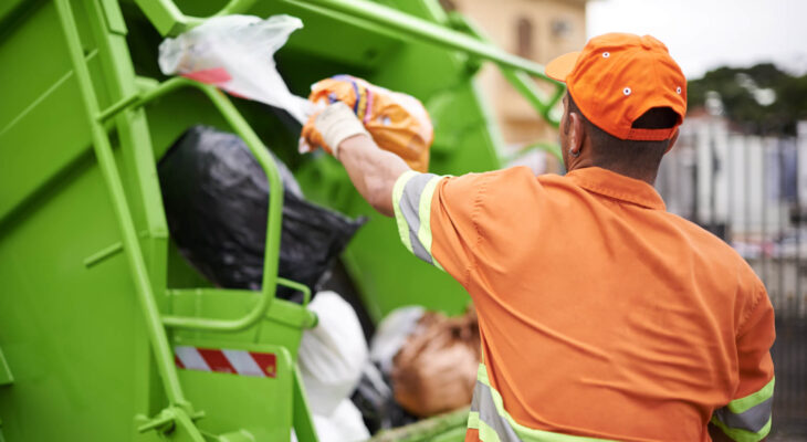 garbage collection worker in front of a green garbage dump truck collecting garbage