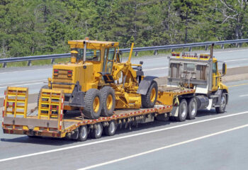 elevated rear quarter view of a flatbed semi truck on a highway transporting a road grader