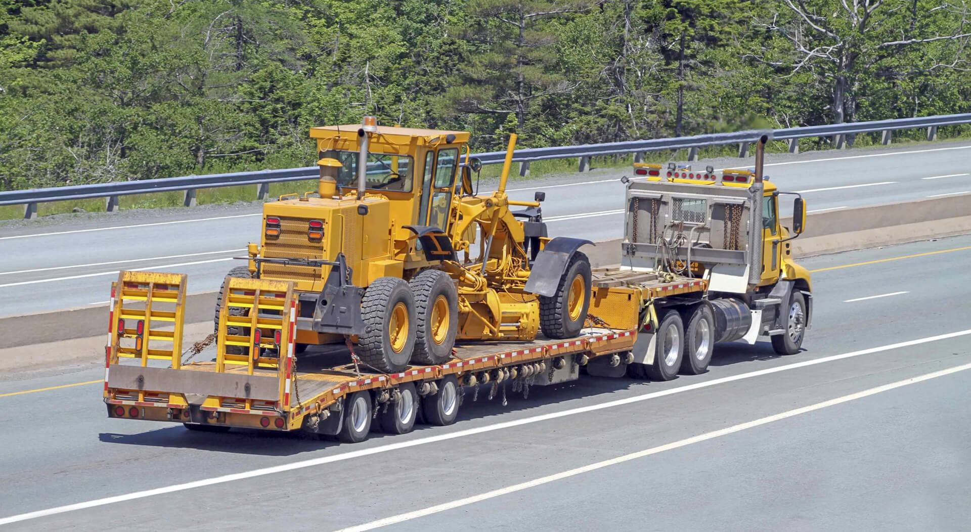 elevated rear quarter view of a flatbed semi truck on a highway transporting a road grader
