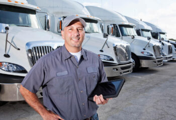 a truck owner is holding a tablet with his left hand while standing in a large parking lot in front of several large trucks