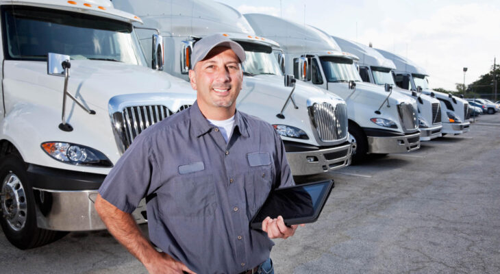 a truck owner is holding a tablet with his left hand while standing in a large parking lot in front of several large trucks