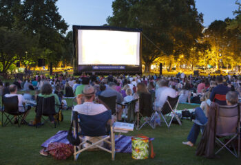 spectators sitting on the grass while watching a movie at an outdoor cinema