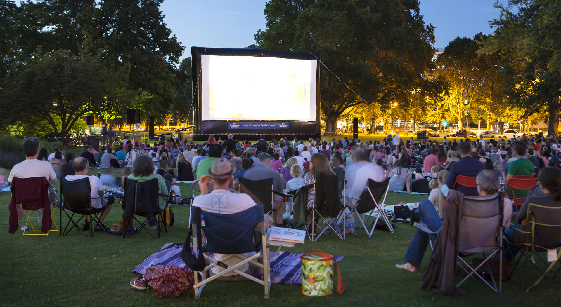 spectators sitting on the grass while watching a movie at an outdoor cinema