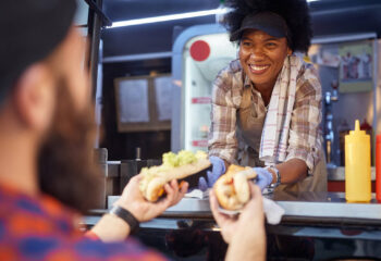polite african-american woman food truck employee giving two sandwiches to a beardy caucasian male customer
