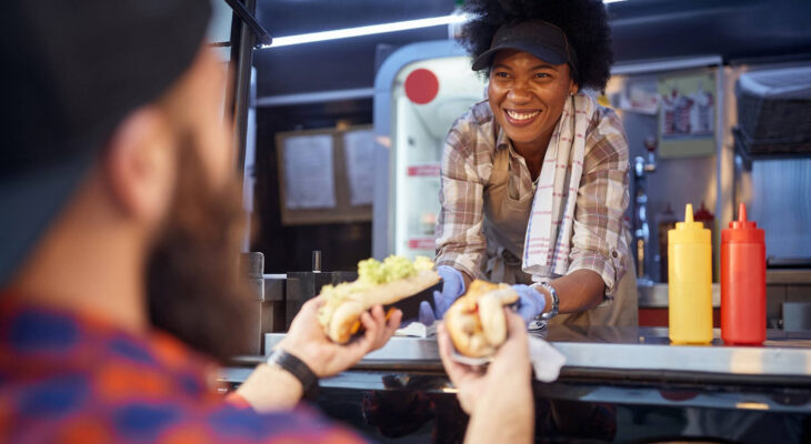 polite african-american woman food truck employee giving two sandwiches to a beardy caucasian male customer