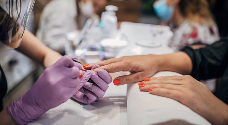 beautician with face shield and mask giving manicure to female customer at beauty salon