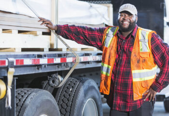 an African-American man in his 30s, standing next to a truck loaded with construction material. He is the truck driver delivering the supplies.