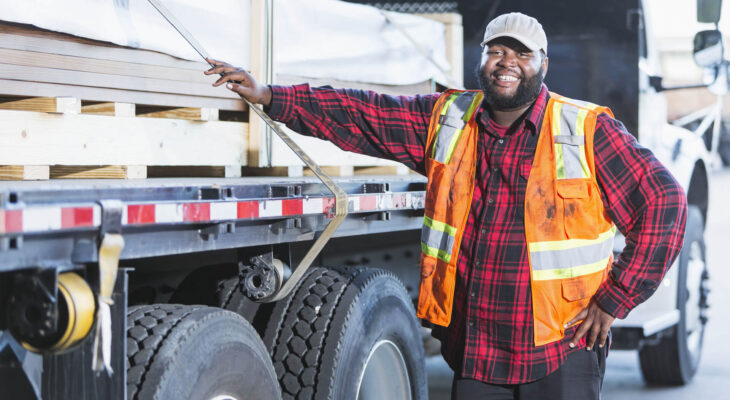 an African-American man in his 30s, standing next to a truck loaded with construction material. He is the truck driver delivering the supplies.