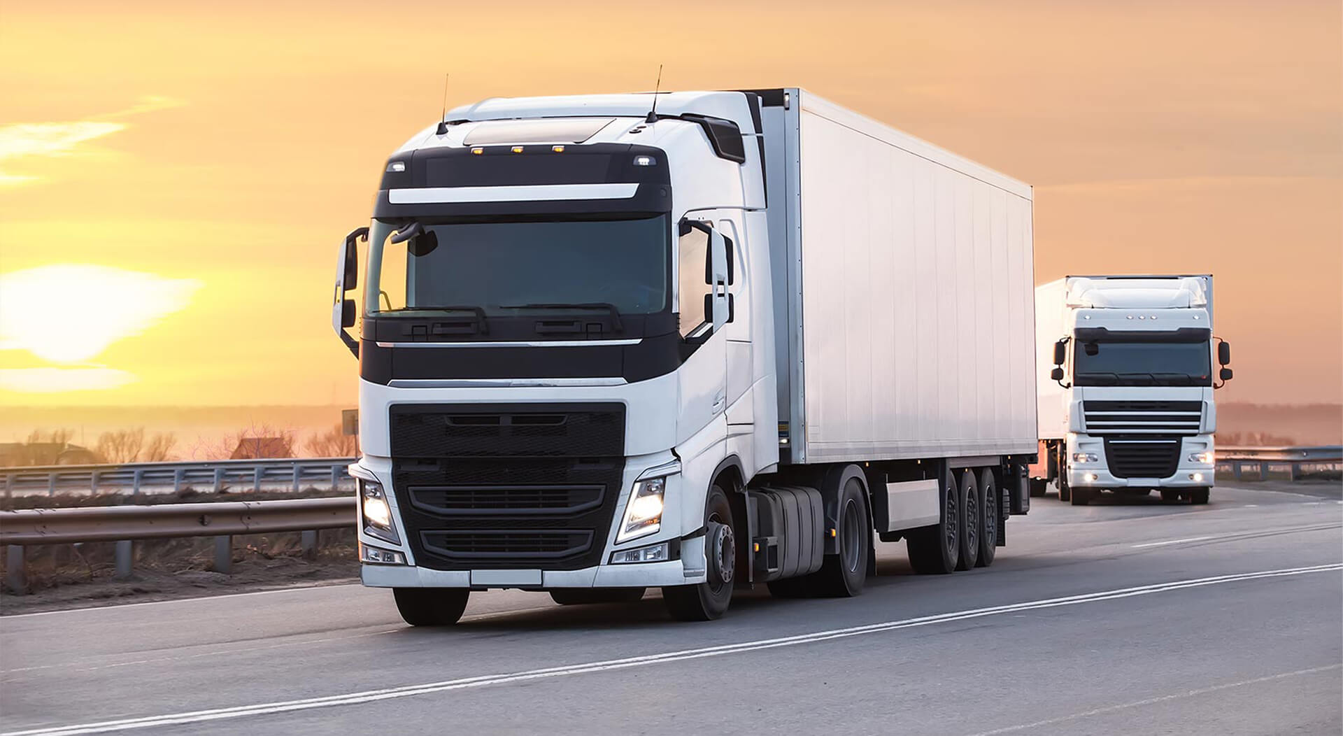 two white high mileage trucks on a highway at sunset