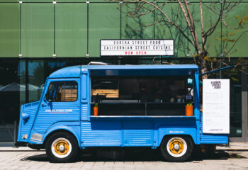 blue food truck parked in front of a green building on a sunny day