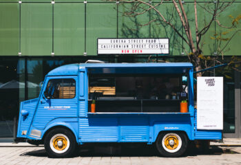 blue food truck parked in front of a green building on a sunny day